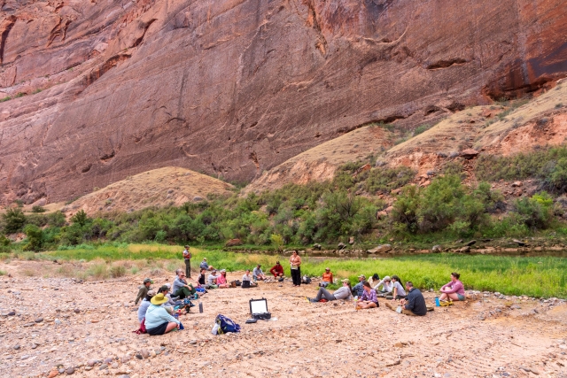 People sit in a circle on the rocky ground with tall canyon walls behind them