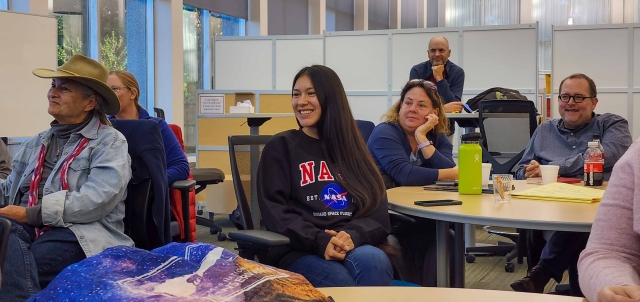 Half a dozen adults seated in a bright meeting area look toward the front of the room while smiling. The woman in the center of the photo wears a NASA sweatshirt.
