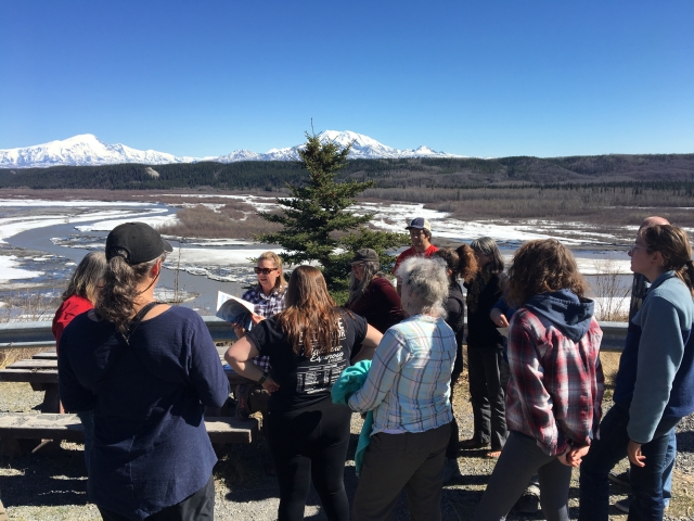 A group of adults gathers around an instructor overlooking an icy river with mountains in the background