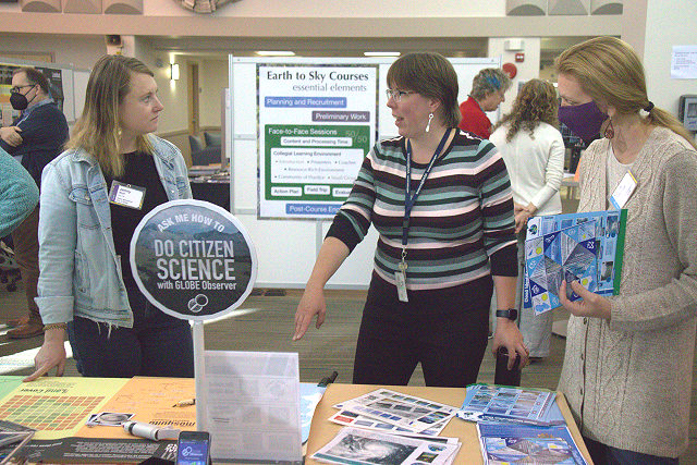 Three women stand around a table. The one on the left wears a jean jacket, the one in the middle wears a striped shirt, and is pointing at the table with printed materials and handouts on it, and the person on the right is wearing a grey sweater and holds a paper. 