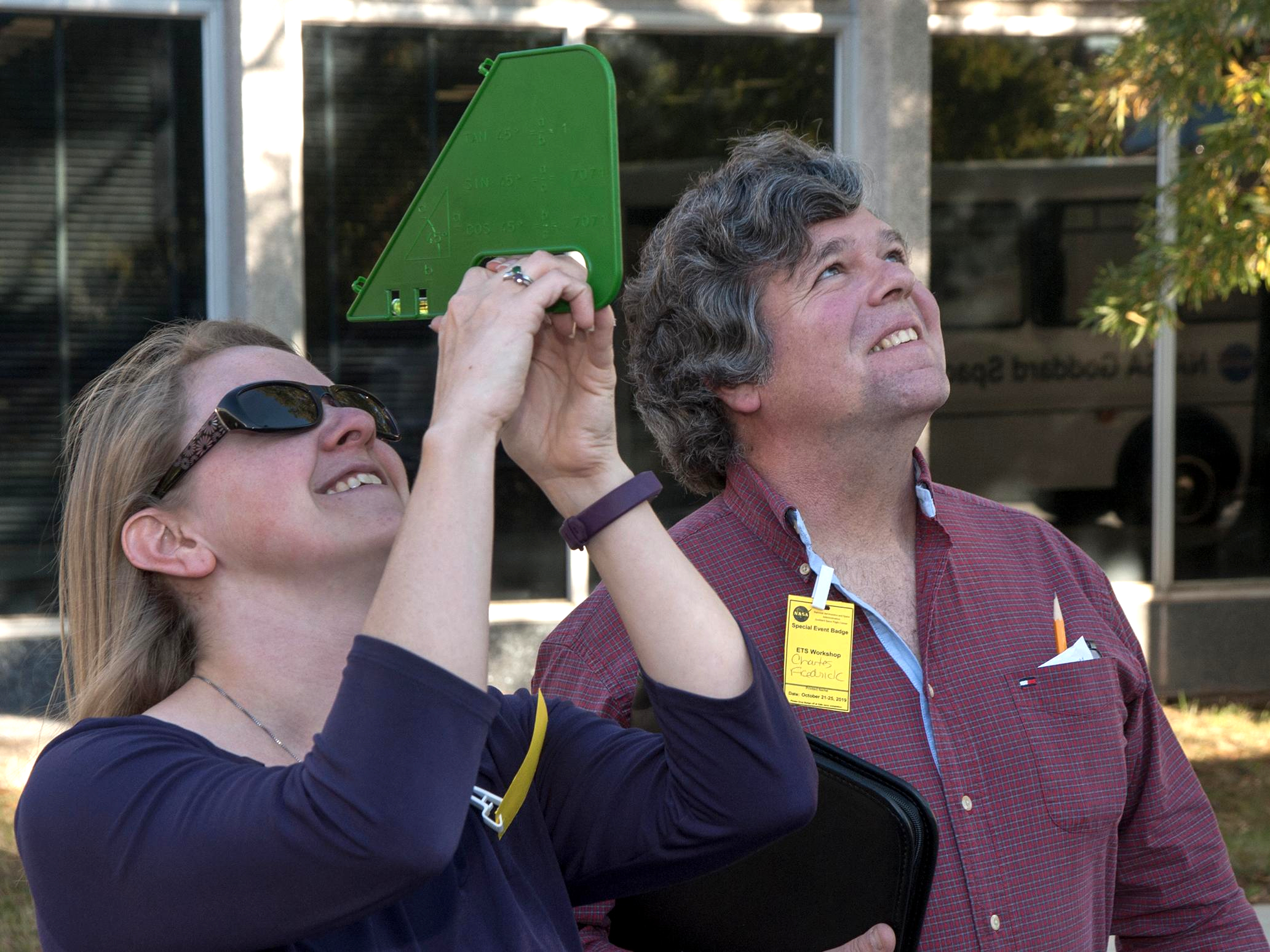 Two people look upwards outdoors using a tool to measure angle