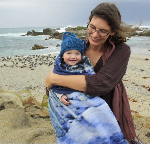A woman with brown hair holding a child wrapped in a blue blanket on a cloudy beach